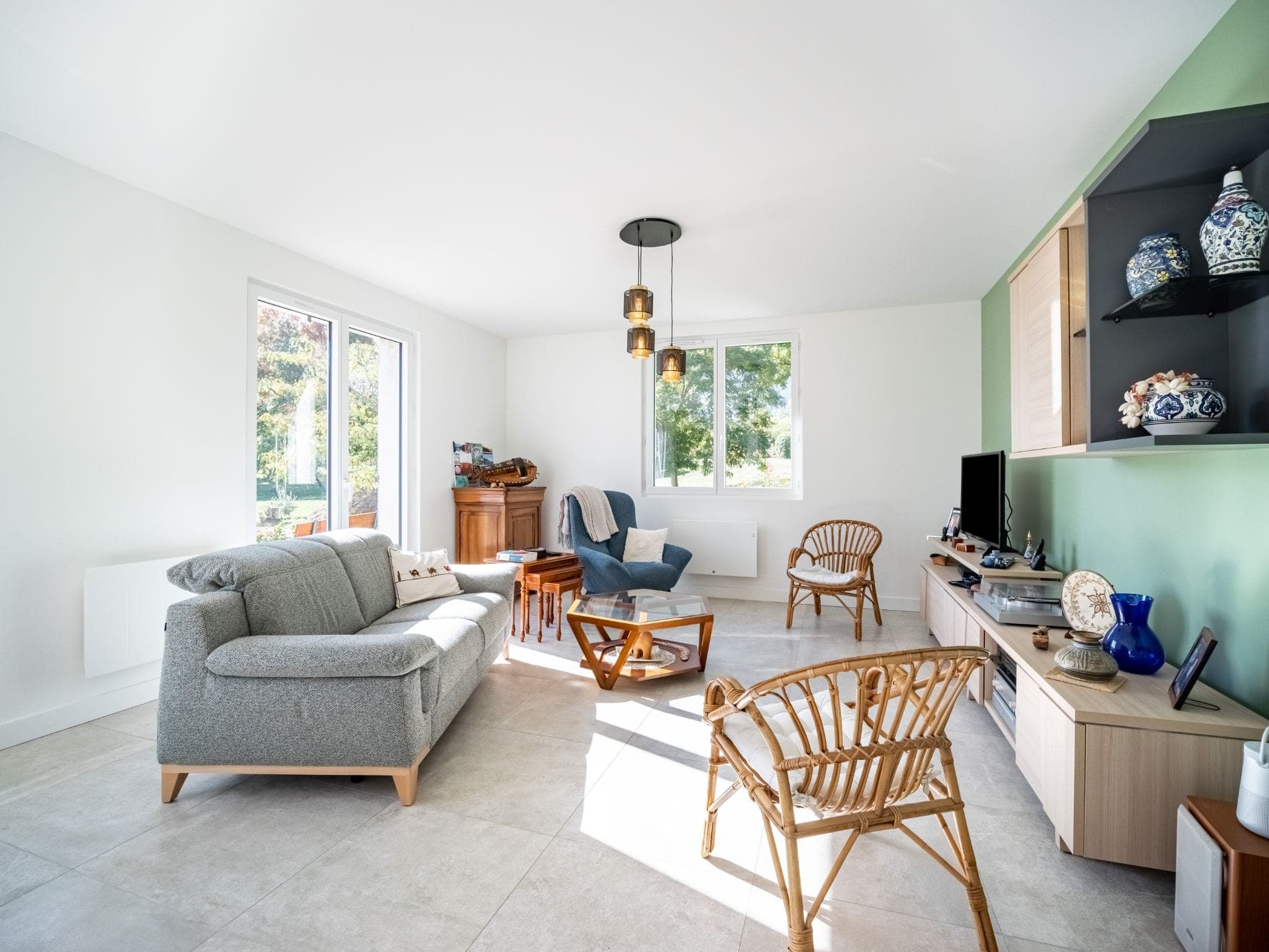 Salon lumineux avec mur vert d’eau, étagères noires et fauteuils en rotin