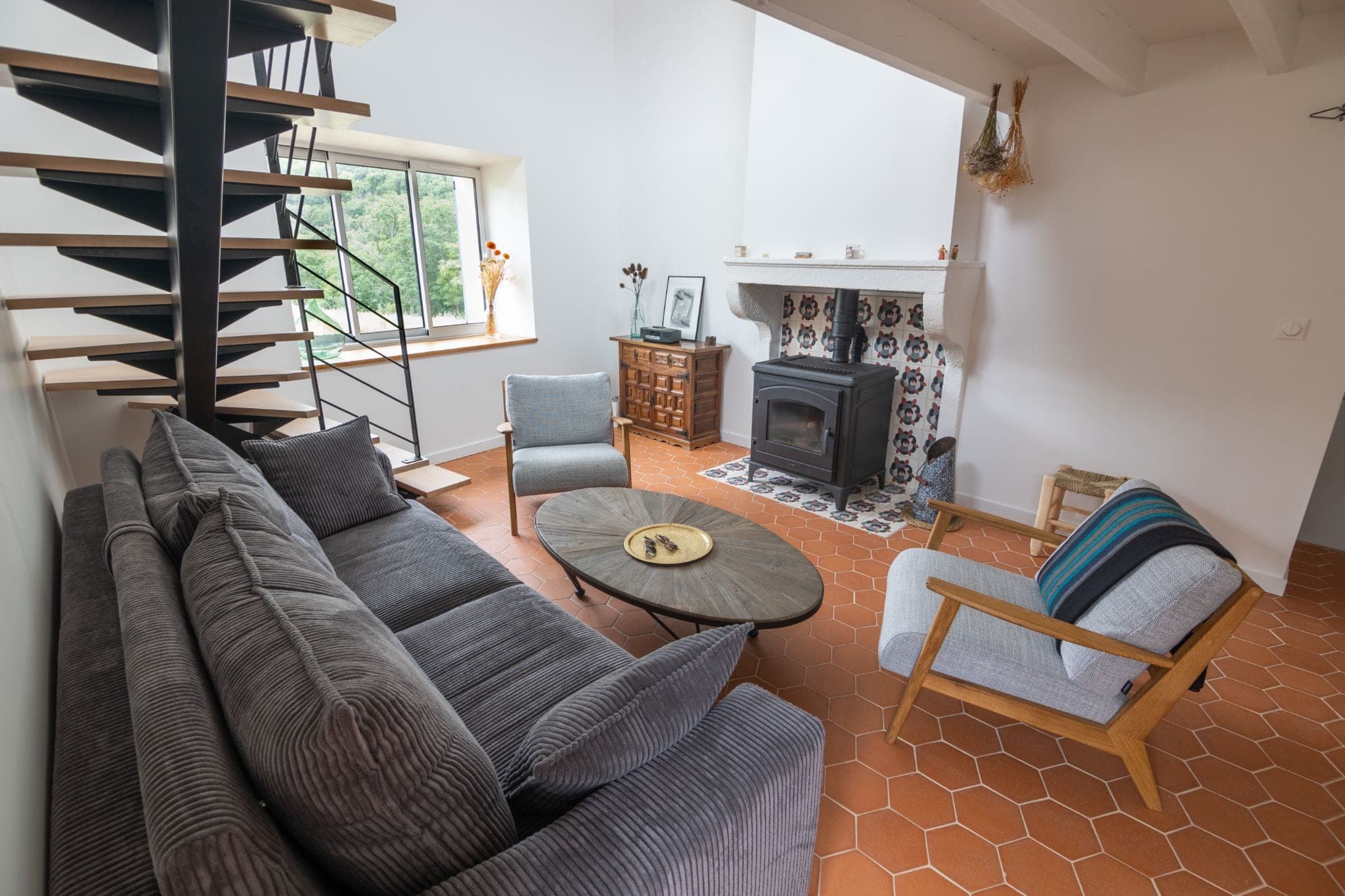 Séjour rénové avec escalier métallique, canapé gris, poêle à bois et tomettes hexagonales